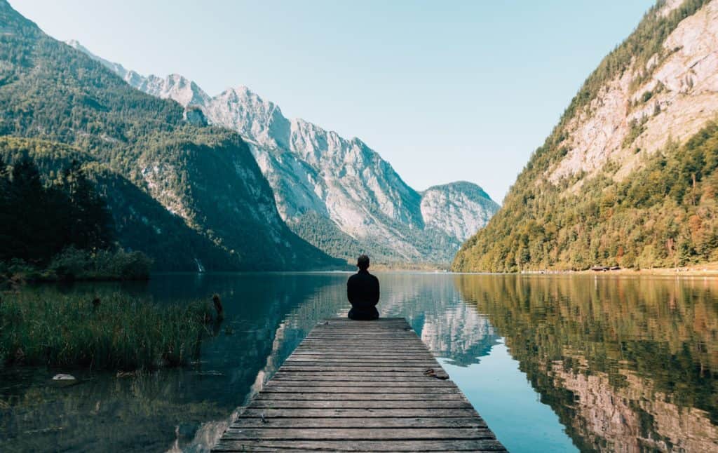 travel photo of a wooden pier with a person sitting and looking out over a river and mountains